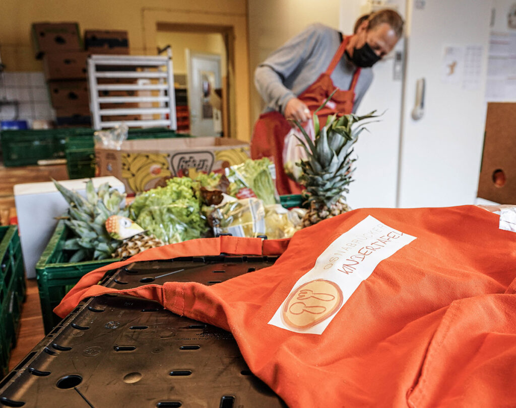 Schürze der Osnabrücker Kindertafel im Vordergrund und im Hintergrund Helfer beim Auspacken von Lebensmitteln